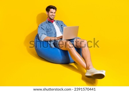 Full size photo of intelligent guy wear jeans jacket shorts sit on pouf chatting on laptop isolated on bright yellow color background