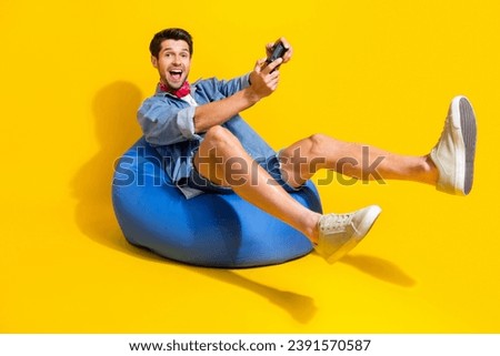 Photo of funky positive guy wear jeans jacket shorts sit on pouf hold playstration joystick isolated on bright yellow color background Royalty-Free Stock Photo #2391570587