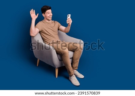 Full size photo of pleasant man dressed beige t-shirt sit in armchair say hi on smartphone video call isolated on blue color background