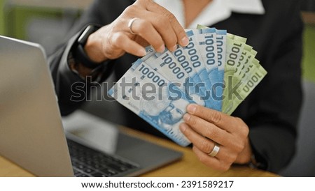 Young beautiful hispanic woman business worker holding chilean pesos using laptop at office