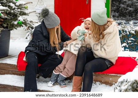 Three pretty young girls sisters celebrating Christmas, sitting on the outdoor terrace with snowing and Christmas decor and Christmas tree and having fun together. celebration, holidays concept.