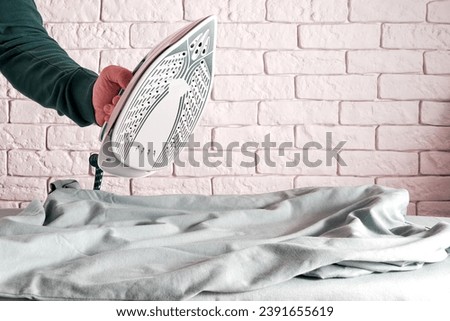 Mans hand holding modern iron over the ironing board and wrinkled t-shirt. Dressing and clothes theme