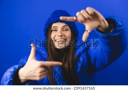 Attractive woman with a lovely smile making a frame gesture with her fingers framing her face. Woman posing on selfie photo looking at camera standing isolated blue background. Girl wear puffer, hat.