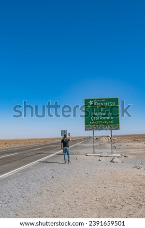 Latin man with long hair, black t-shirt and jeans photographing the Tropic of Capricorn signpost in the Atacama Desert, Chile.