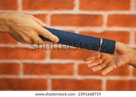 Diploma holder, in dark blue color, with gold details, being held by a female hand with light skin, handed to another male hand with light skin, crosswise. Blurred brick wall background.