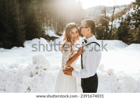 Portrait of the bride and groom against the background of a pine forest and backlight. The bride in a white wedding dress, the groom in a white shirt and trousers with suspenders. Winter wedding.