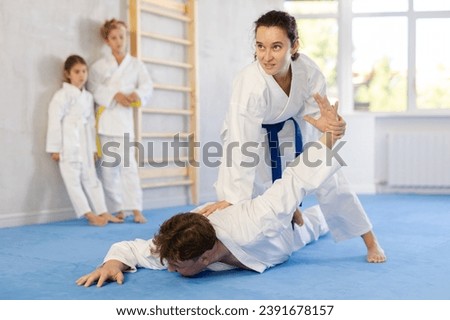 Concentrated young woman attending family karate class, applying armlock technique to husband while preteen children in kimonos watching training sparring in background Royalty-Free Stock Photo #2391678157