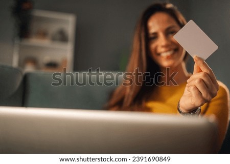 Close up of hand holding a credit card and showing it at the camera with a blurry woman in a background. A woman is holding a credit card and smiling while using technologies for e-banking.
