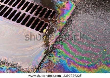 An oil slick against the backdrop of an asphalt road flows into a storm drain through a grate. Royalty-Free Stock Photo #2391729225