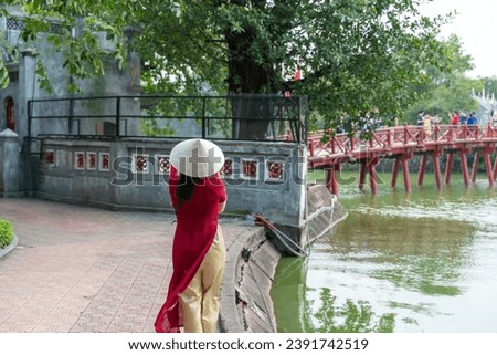 Female tourist wearing Vietnamese traditional dress Ao Dai with The Huc bridge at Hoang Kiem lake on background Royalty-Free Stock Photo #2391742519