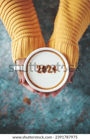 Happy New Year 2024 theme number 2024 on frothy surface of cappuccino served in white coffee mug holding by female hands over rustic blue background. Holidays food art, new year new you. (top view) Royalty-Free Stock Photo #2391787975
