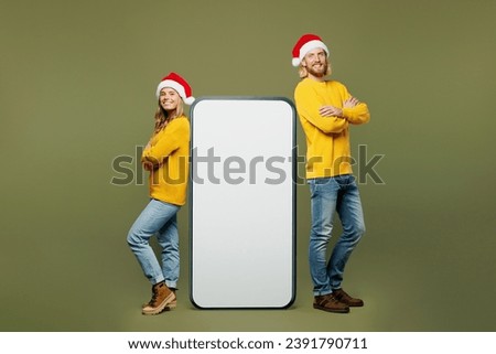 Full body merry young couple two friends man woman wear sweater Santa hat posing stand near big huge blank screen mobile cell phone isolated on plain green background. Happy New Year Christmas concept