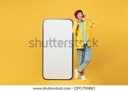 Full body young woman wears raincoat red hat big huge blank screen mobile cell phone listen music in headphones isolated on plain yellow background. Outdoors lifestyle wet fall weather season concept