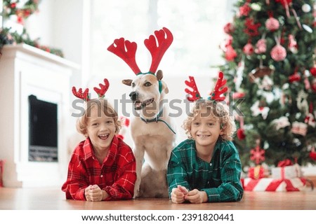 Christmas at home. Kids and dog in reindeer antlers under Xmas tree. Little child hug puppy in deer hat and open Christmas presents. Children play with pet. Winter holiday celebration. 