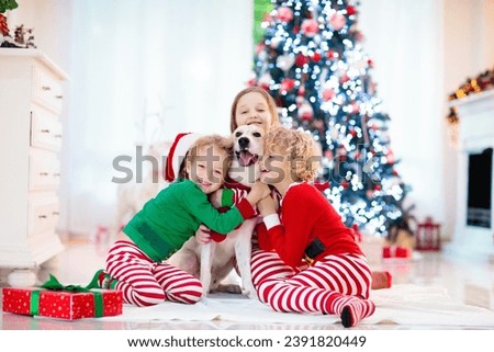 Christmas at home. Kids and dog under Xmas tree. Little boy and girl hug pet in Santa hat and open Christmas presents. Children play with animal. Winter holiday celebration. Love and friendship.
