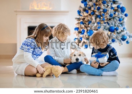 Christmas at home. Kids and dog under Xmas tree. Little boy and girl hug pet in Santa hat and open Christmas presents. Children play with animal. Winter holiday celebration. Blue and white theme.