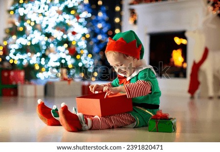 Child opening present at Christmas tree at home. Kid in elf costume with Xmas gifts and toys. Little baby boy with gift box and candy at fireplace. Family celebrating winter holidays. Home decoration.