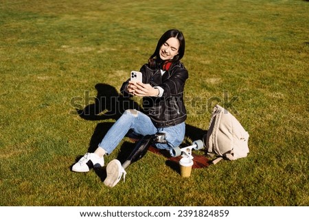 Young woman student with prosthetic leg sitting on green grass in university campus and making selfie with smartphone. Disabled woman with bionic leg. Woman with leg prosthesis equipment using phone.