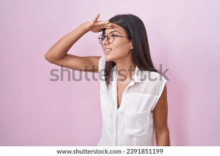 Brunette young woman standing over pink background wearing glasses very happy and smiling looking far away with hand over head. searching concept. 