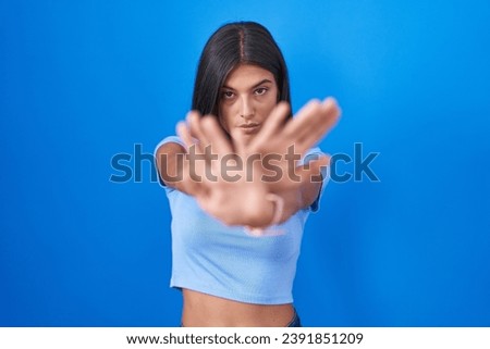 Brunette young woman standing over blue background rejection expression crossing arms and palms doing negative sign, angry face 