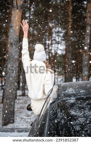 Young woman in woolen white hat have a fun in black car. Winter travel, snow-covered trees in the forest. Snowy weather. High quality photo