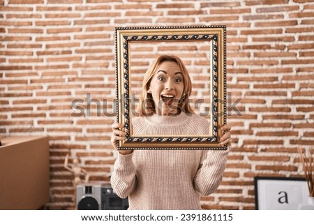 Hispanic woman holding empty frame celebrating crazy and amazed for success with open eyes screaming excited. 