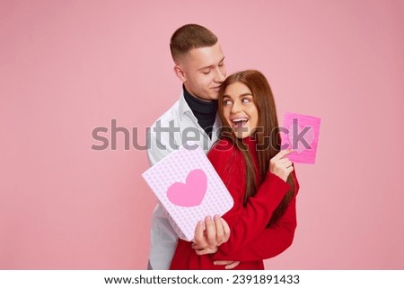 Greetings. Beautiful young couple, man and woman hugging, holding postcard with hearts against pink studio background. Concept of love, relationship, Valentine's Day, emotions, lifestyle