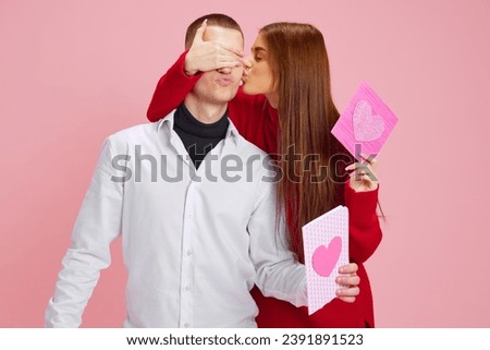 Kiss. Young girl covering boyfriend's eyes and presenting him little gift, postcard with heart against pink studio background. Concept of love, relationship, Valentine's Day, emotions, lifestyle