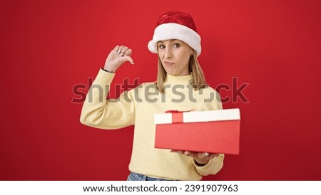 Young blonde woman unpacking christmas gift looking upset over isolated red background