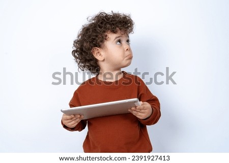 Beautiful two year old toddler with curly hair wearing casual clothes over white studio background holding a tablet pad and looking aside with sad face