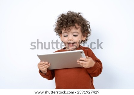 Beautiful two year old toddler with curly hair wearing casual clothes over white studio background holding a tablet pad and smiling