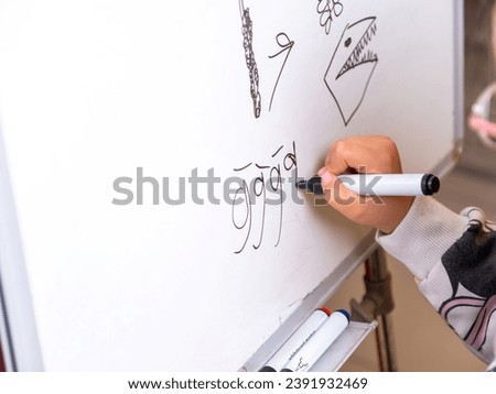 School girl trying to draw the letter g on the whiteboard. School girl learning how to draw. Close-up of school girl's hands drawing on white board.   Royalty-Free Stock Photo #2391932469