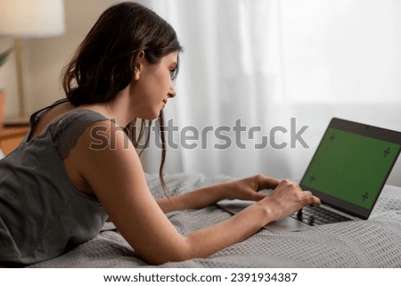 Indoor shot of young woman using laptop with green screen. Brunette girl lying on the bed and typing on computer keyboard with chroma key