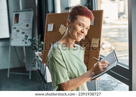 stylish queer manager smiling at camera and writing in notebooks in modern office environment