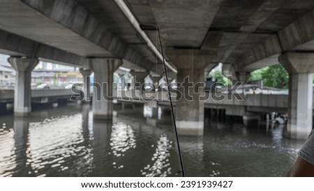 Image of a person using a small fishing rod. Fishing is in the area of ​​the waterway passing under the elevated road. In the picture you can see the path under the bridge. with cars passing by
