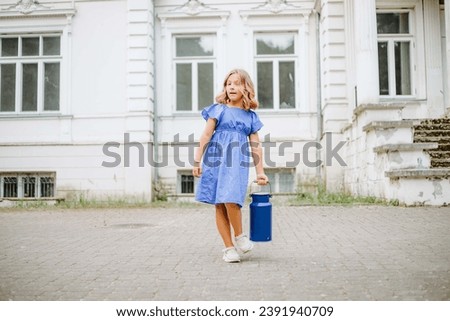 Lovely girl with long blond hair in bright blue dress with blue antique can stands against background of classic white house. Beautiful girl 8-10 years old. Carefree childhood. Children of model . Royalty-Free Stock Photo #2391940709
