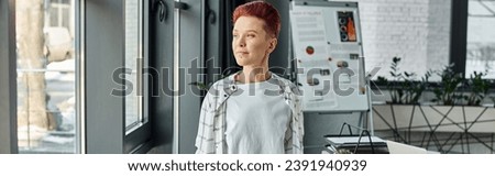 thoughtful queer person in casual attire standing near windows and looking away in office, banner
