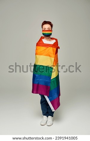 full length of redhead queer person in rainbow colors medical mask posing in LGBT flag on grey