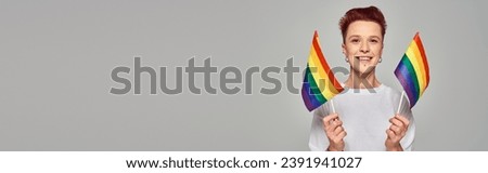 joyful queer person in white t-shirt holding small LGBT flags and looking at camera on grey, banner