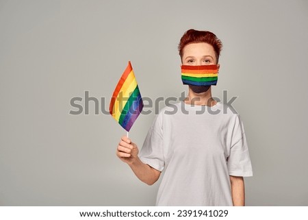 redhead queer person in white t-shirt and rainbow colors medical mask with small LGBT flag on grey