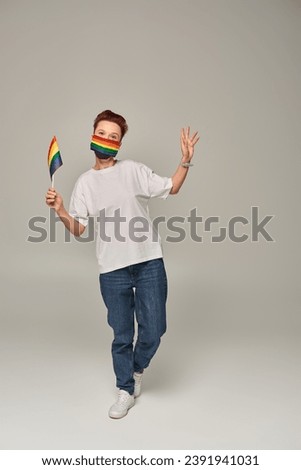 full length of redhead queer person in rainbow colors medical mask with small LGBT flag on grey