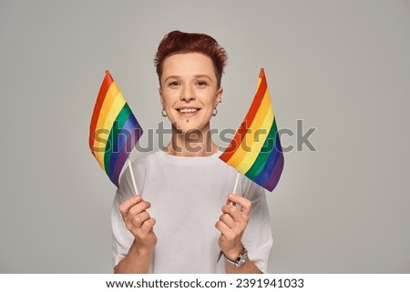 joyful queer person in white t-shirt holding small LGBT flags and looking at camera on grey backdrop