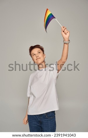 redhead queer person in white t-shirt holding small LGBT flag in raised hand while standing on grey