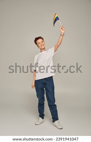 cheerful queer person in white t-shirt holding small LGBT flag in raised hand while standing on grey