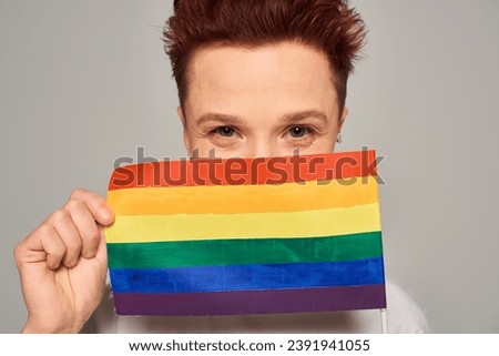 joyful redhead queer person obscuring face with small LGBT flag and looking at camera on grey