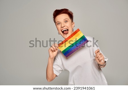 excited queer model with piercing holding small LGBT flat and sticking out tongue on grey backdrop