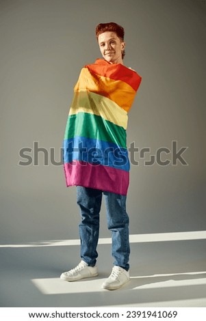 full length of smiling queer person posing with rainbow colors LGBT flag white standing on grey