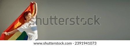 happy and unique queer person in white t-shirt posing with rainbow colors LGBT flag on grey, banner