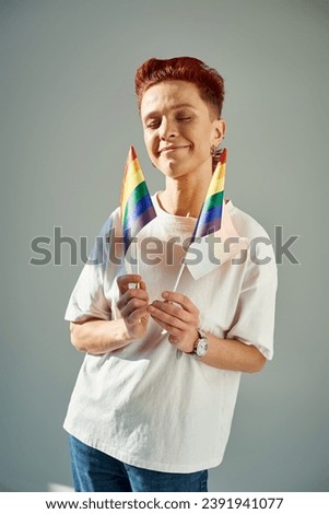 redhead queer person in white t-shirt holding small LGTB flags and smiling with closed eyes on grey