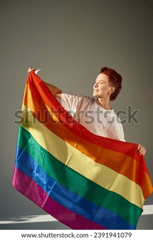 happy and unique queer person in white t-shirt posing with rainbow colors LGBT flag on grey backdrop